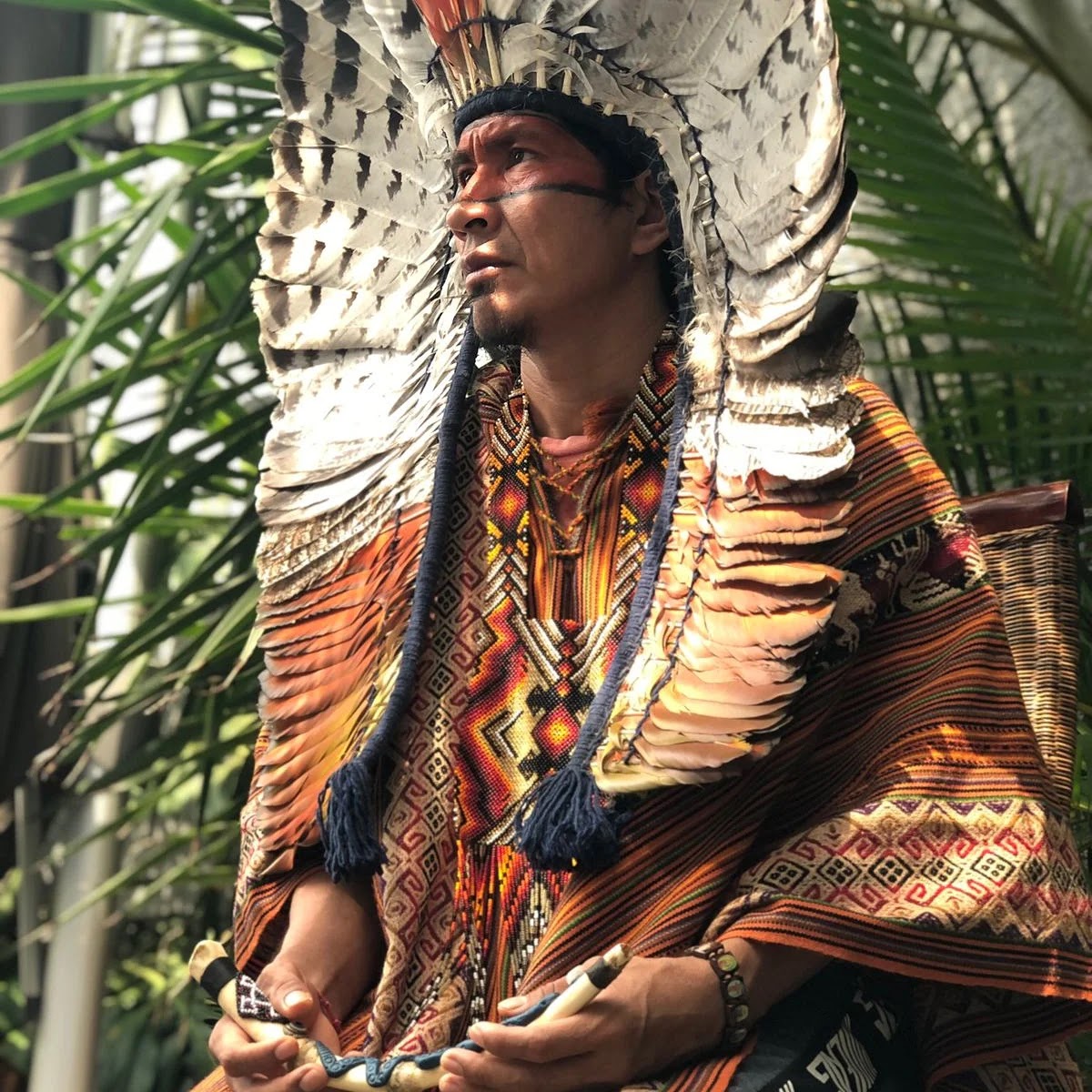 Xinu Yawanawa en costume cérémoniel avec coiffe de plumes blanches rayonnantes, poncho brodé et flûtes cérémonielles — incarnation visuelle de Txana, l'oiseau passeur de culture