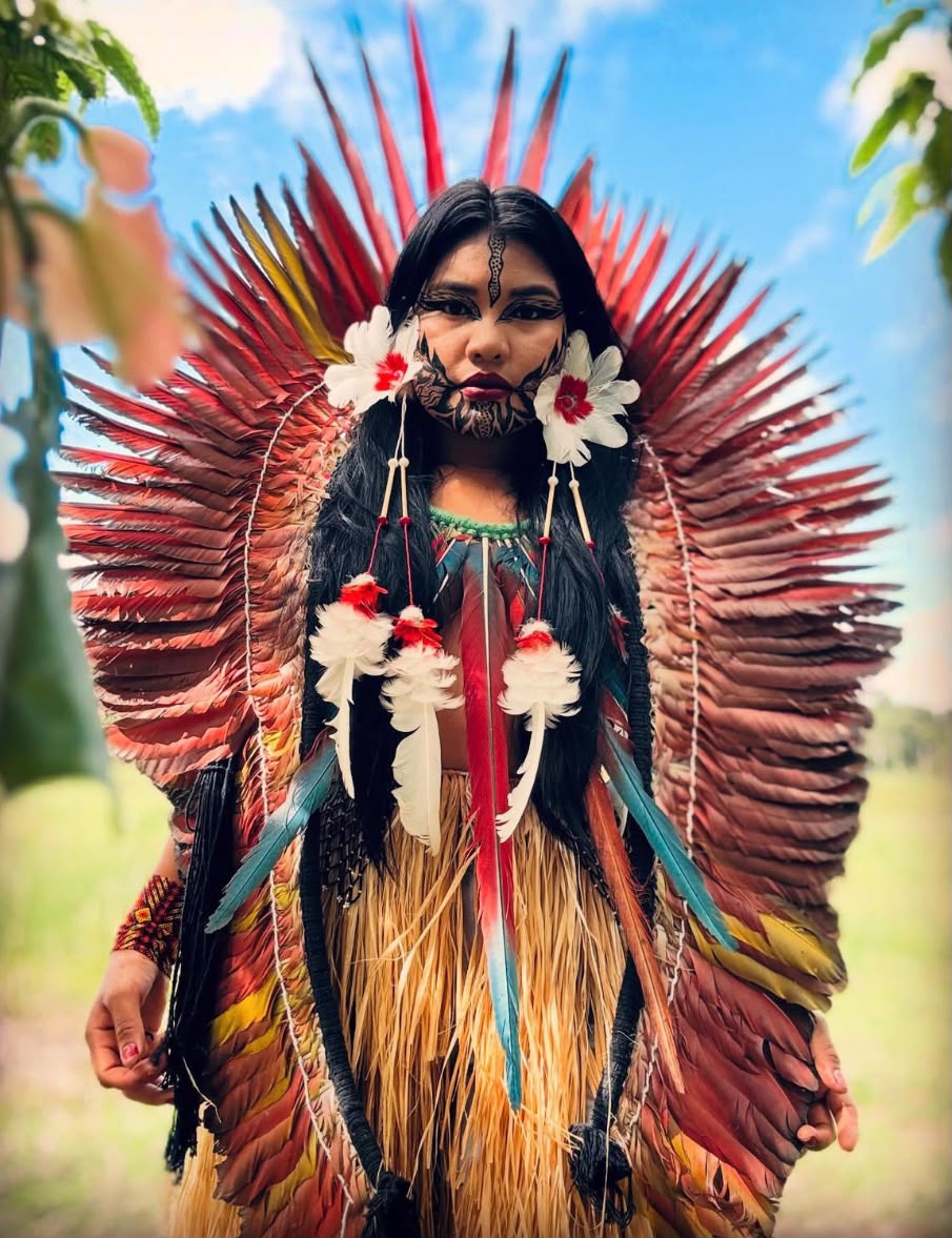 Yaka Yawanawa portant le cocar Txana — coiffe éventail de plumes rouges rayonnantes, peinture faciale rituelle et jupe en fibres naturelles — incarnation vivante de Txana, l'oiseau passeur de culture