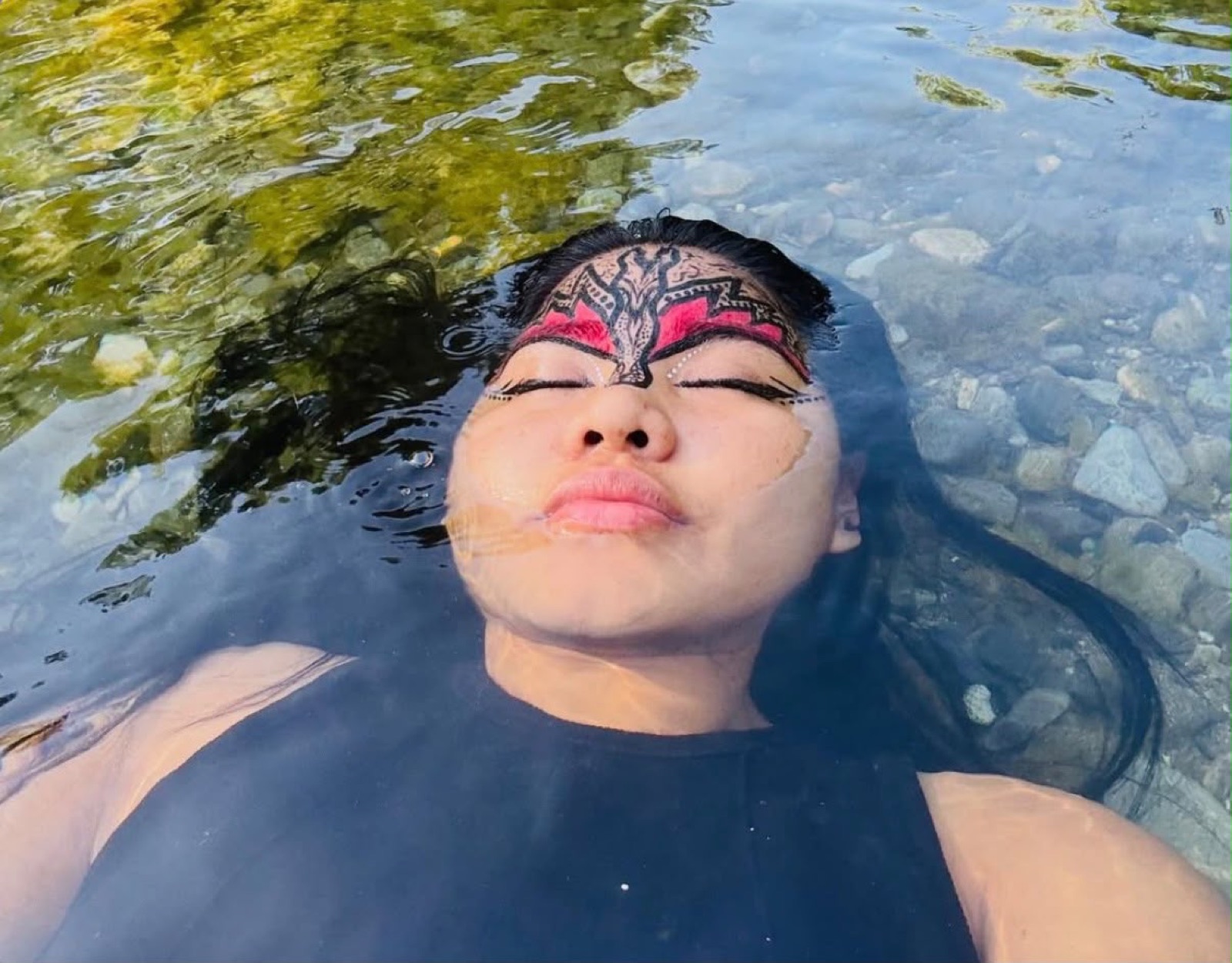 Yaka Yawanawa, visage porteur d'une peinture rituelle rouge et noire, flottant paisiblement à la surface d'une rivière — l'eau, l'ancestralité et le calme réunis